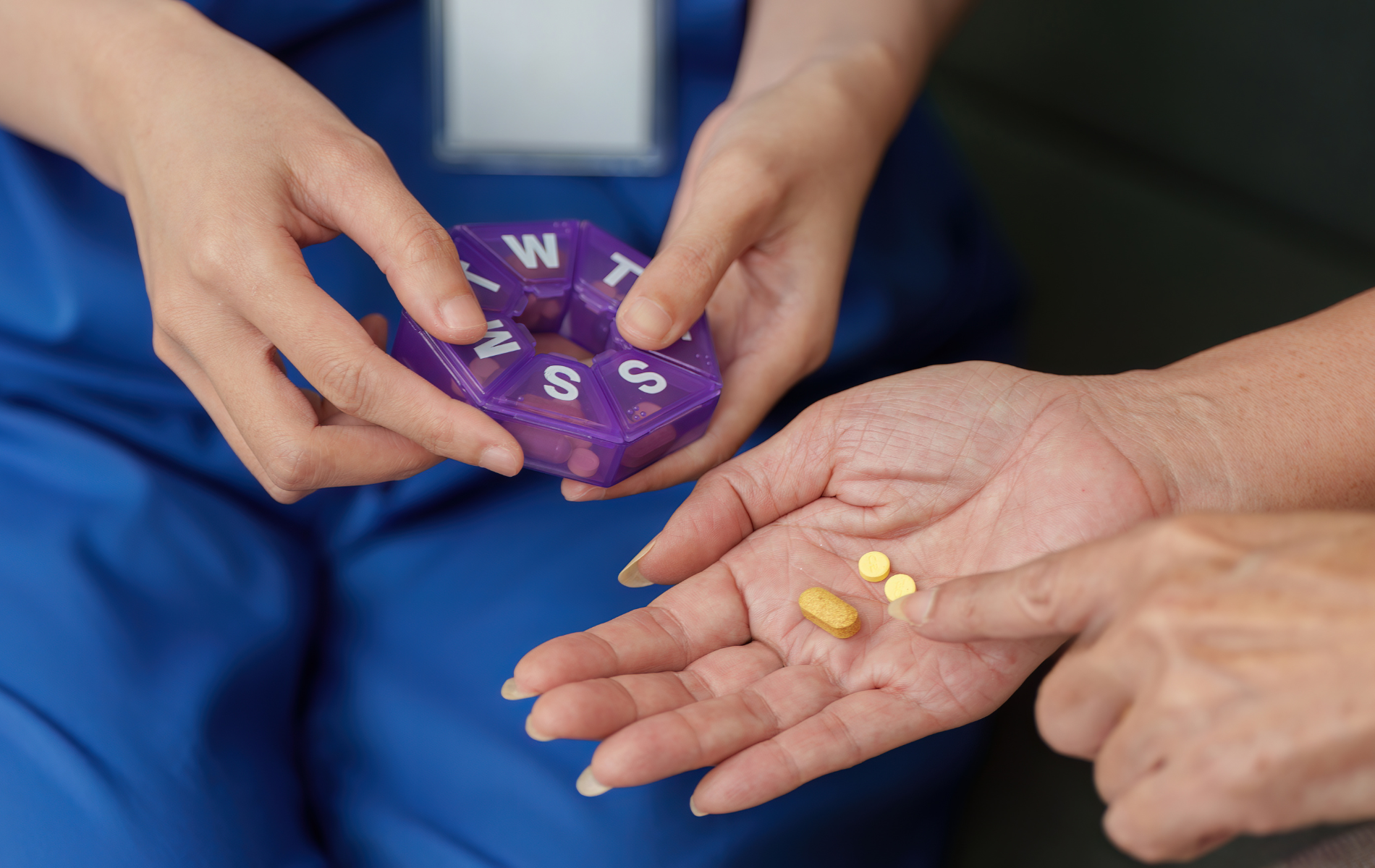 Nurse dispensing pills from a purple pill organizer into a patient's hand.