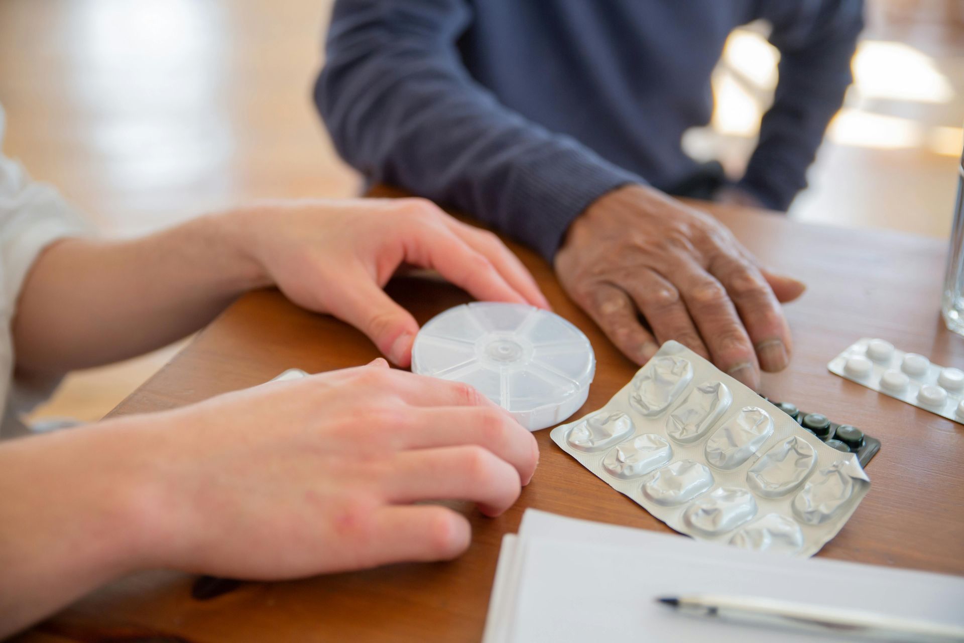 Hands sorting pills and a white pill organizer on a table beside blister packs and paper.