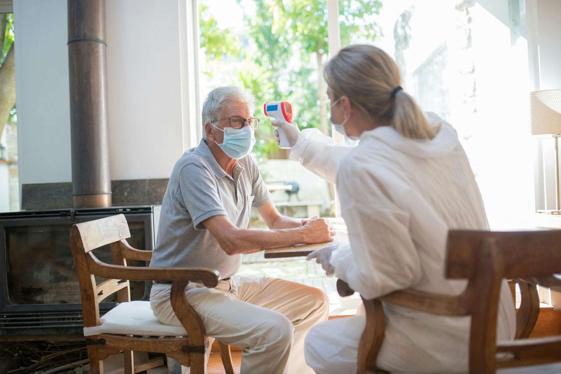 Two masked people talking at a sunlit table in a cozy room