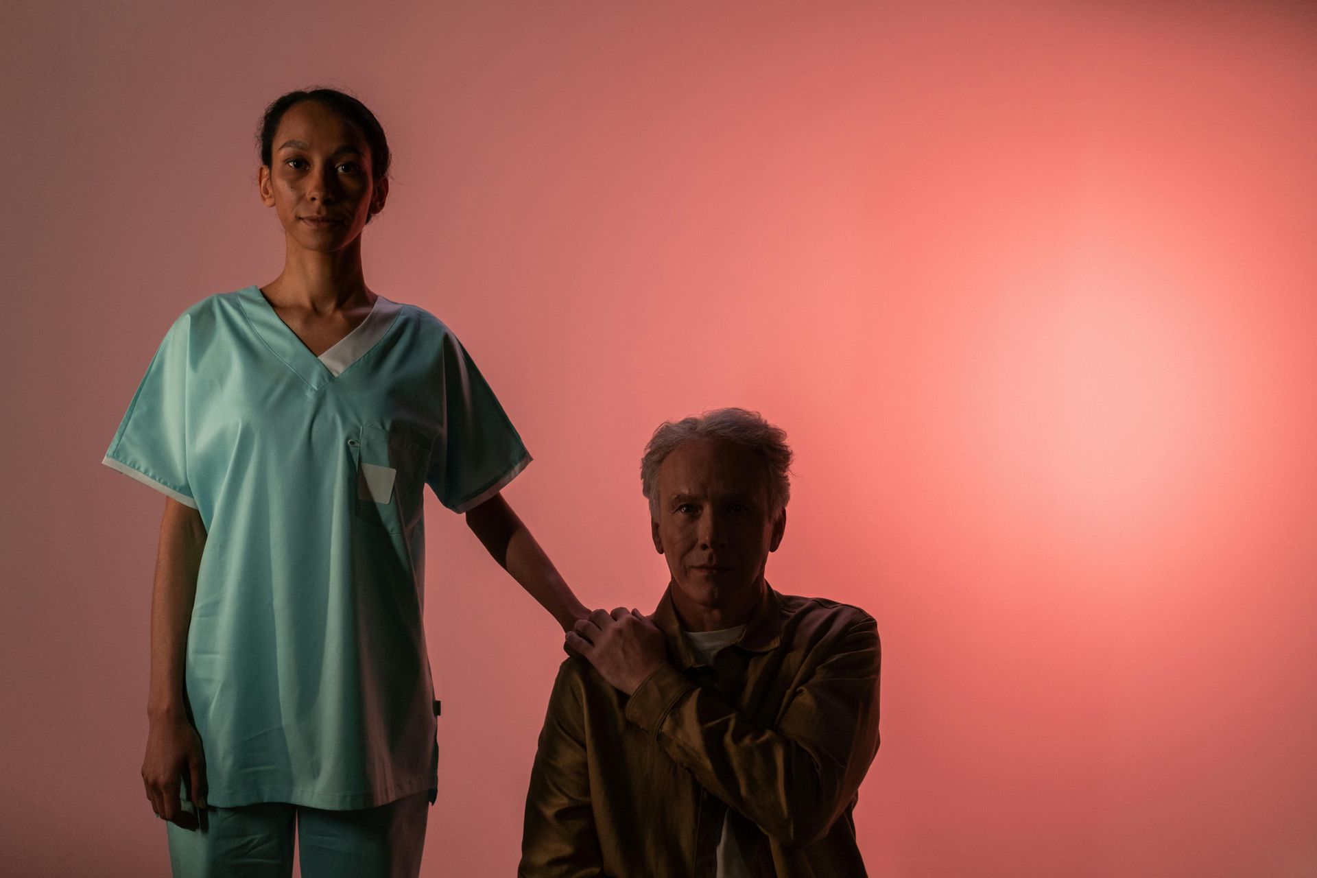 Two people in medical scrubs pose against a pink-lit background, one standing and one seated.