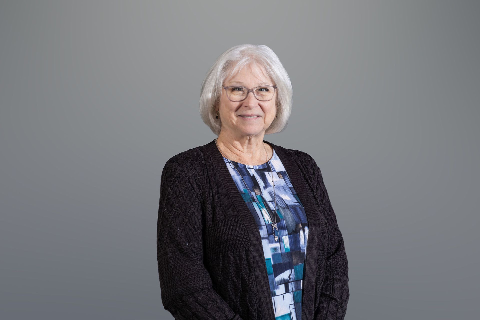 Smiling older woman with white hair and glasses, wearing a black cardigan over a patterned blouse on a gray background
