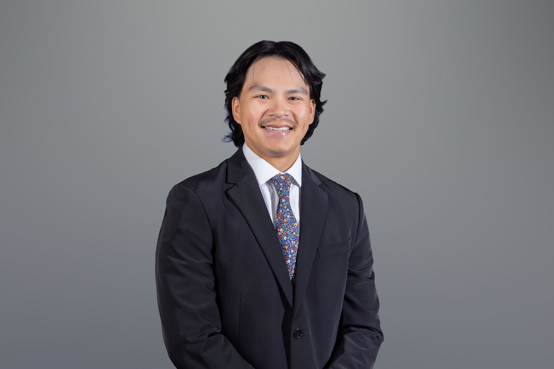 Smiling man in a black suit and patterned tie against a gray background
