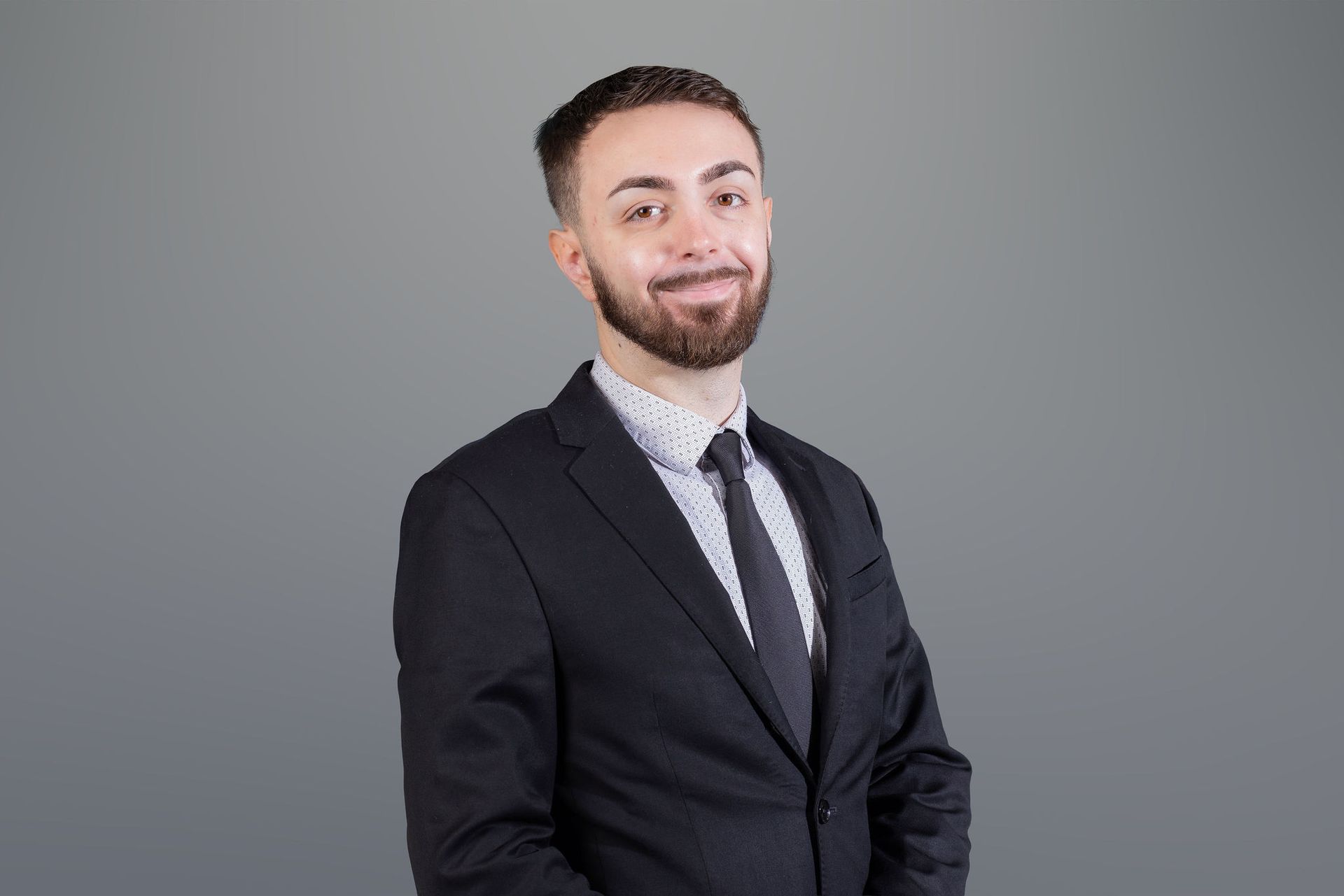 Man in a black suit, white shirt, and tie smiling against a gray studio background
