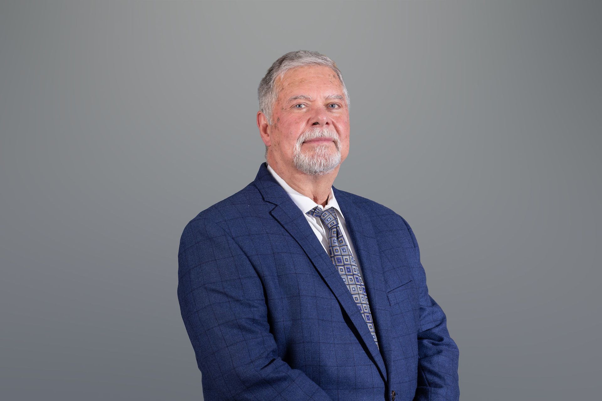 Man in a blue suit with a patterned tie, seated against a plain gray background.