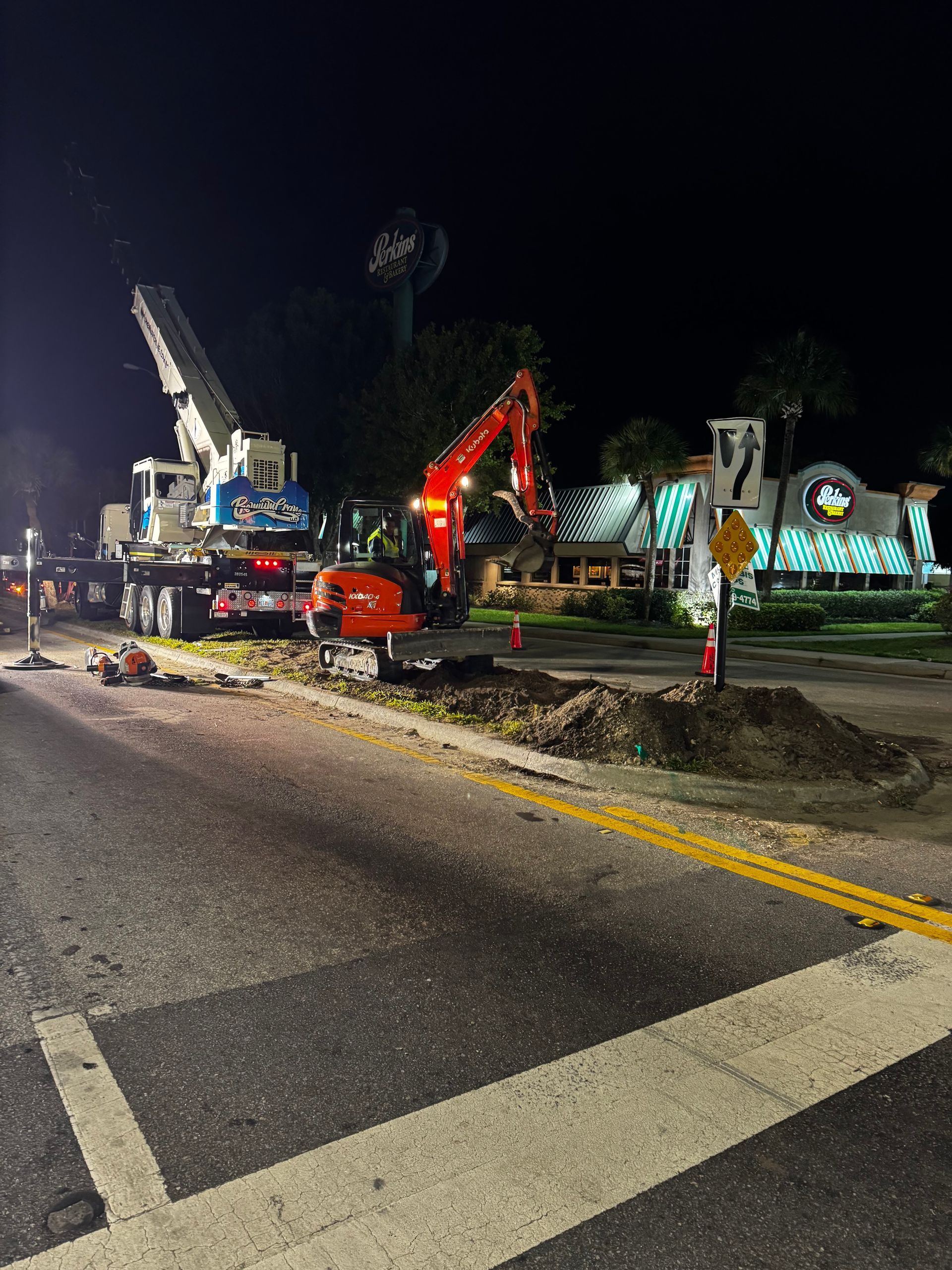 A crane and an excavator are on the side of the road at night.