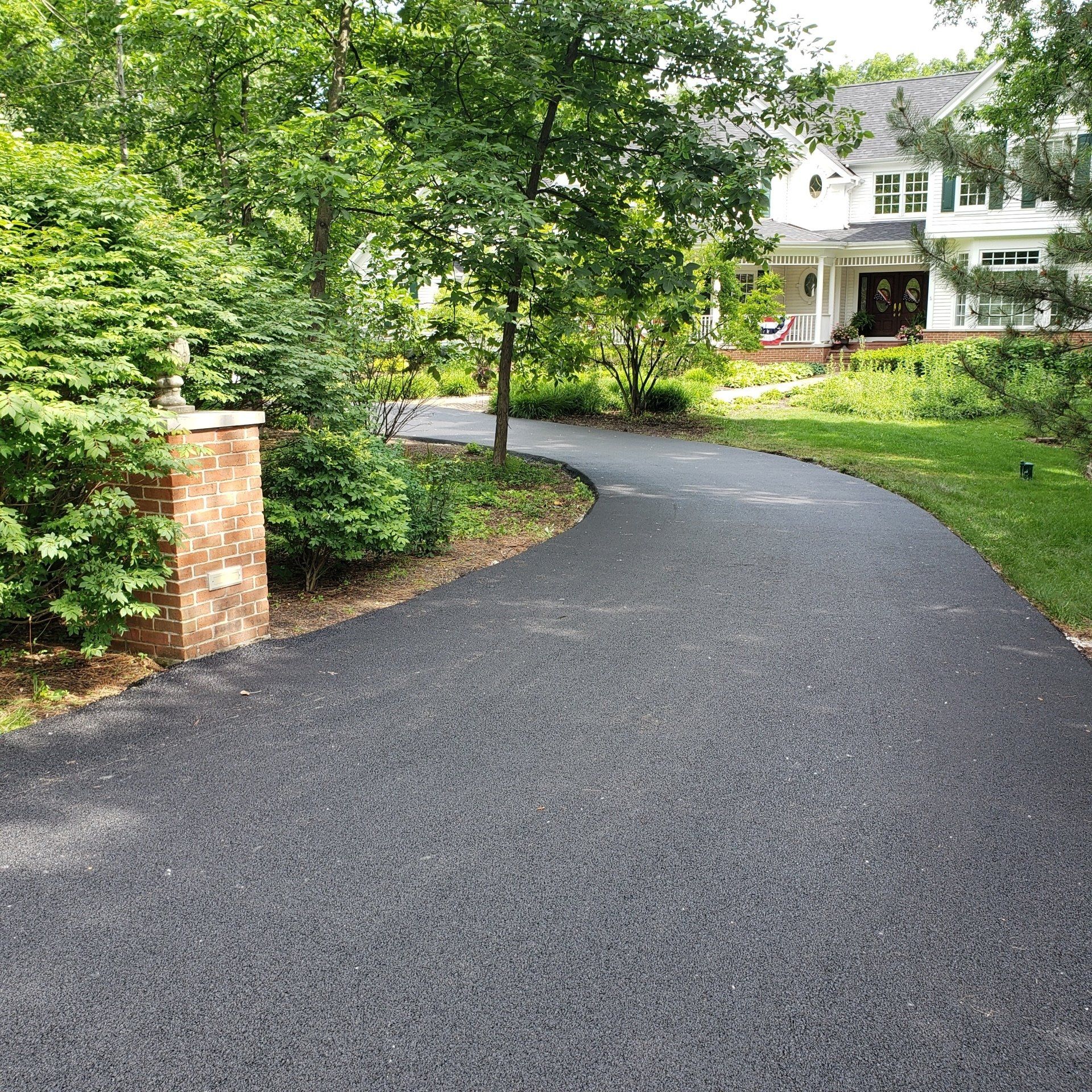 A curved asphalt driveway leads to a white house surrounded by green trees and a brick pillar on the left.