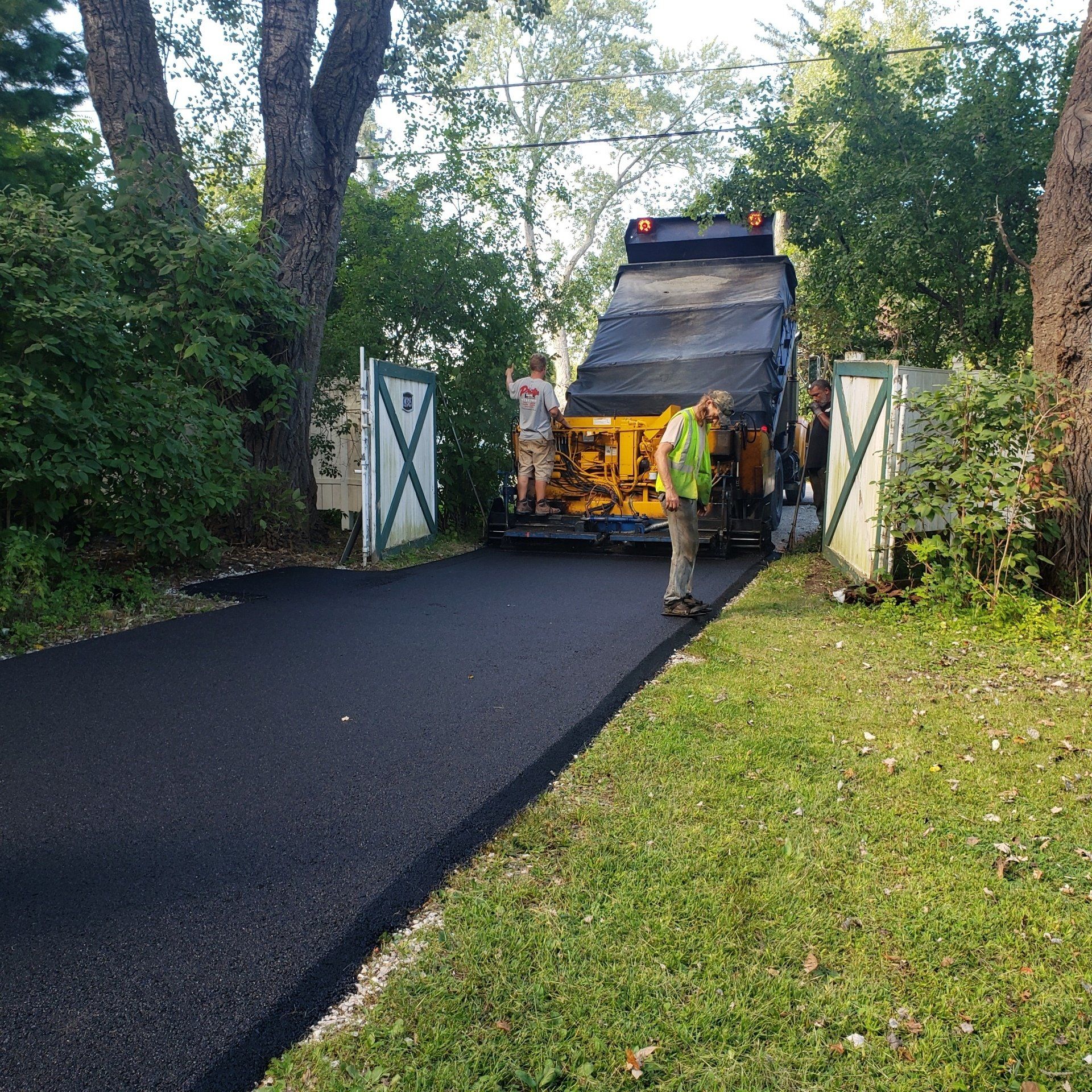 Workers operate a yellow paving machine to lay fresh asphalt on a driveway between two white gates in a wooded area.