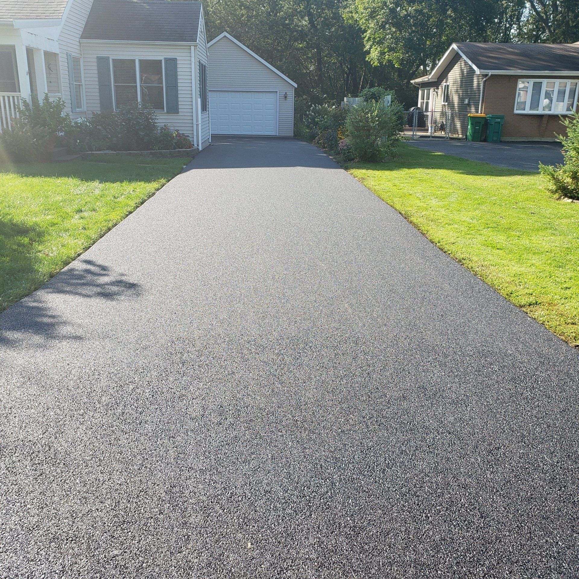 A freshly paved black asphalt driveway leads to a white detached garage between two houses on a sunny day.