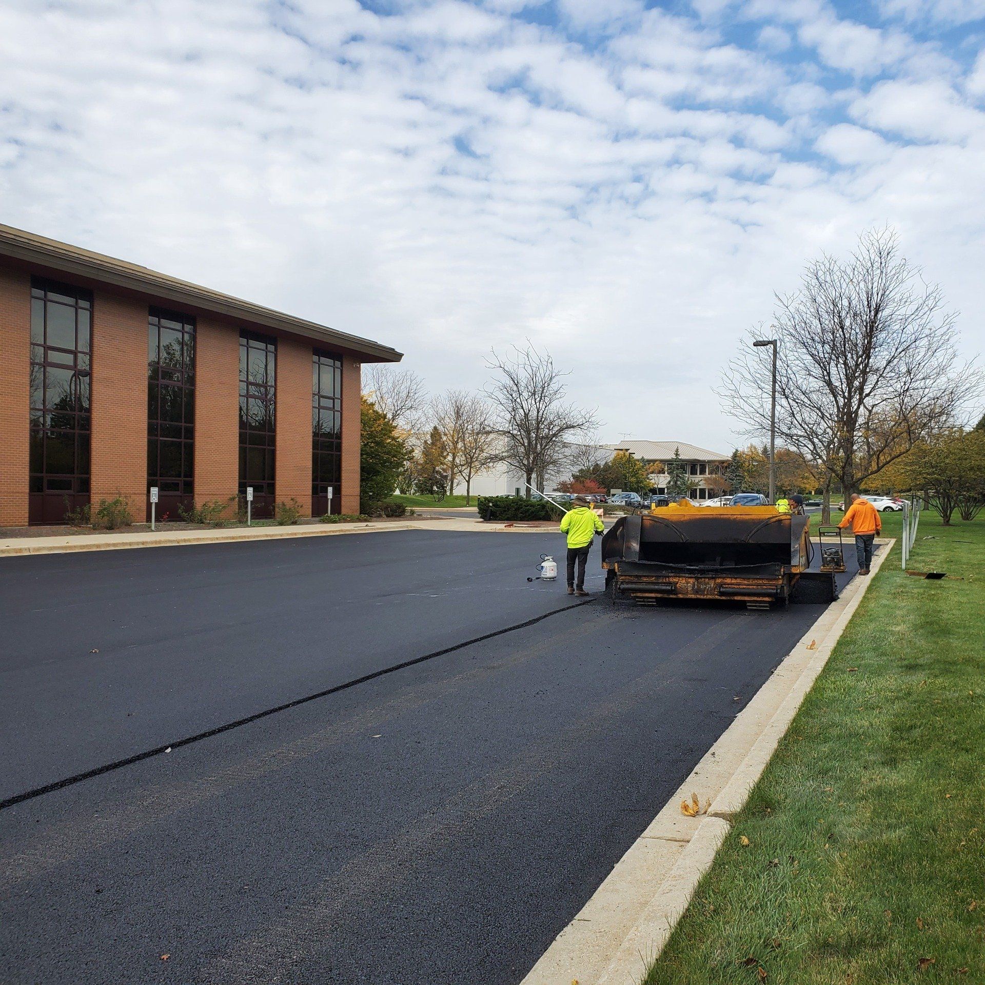 Fallen Tree Blocking Road | Lake Villa, IL | Prestige Paving and Seal Coating Asphalt paving in progress; two workers operate a paving machine next to a building, creating a new black surface.