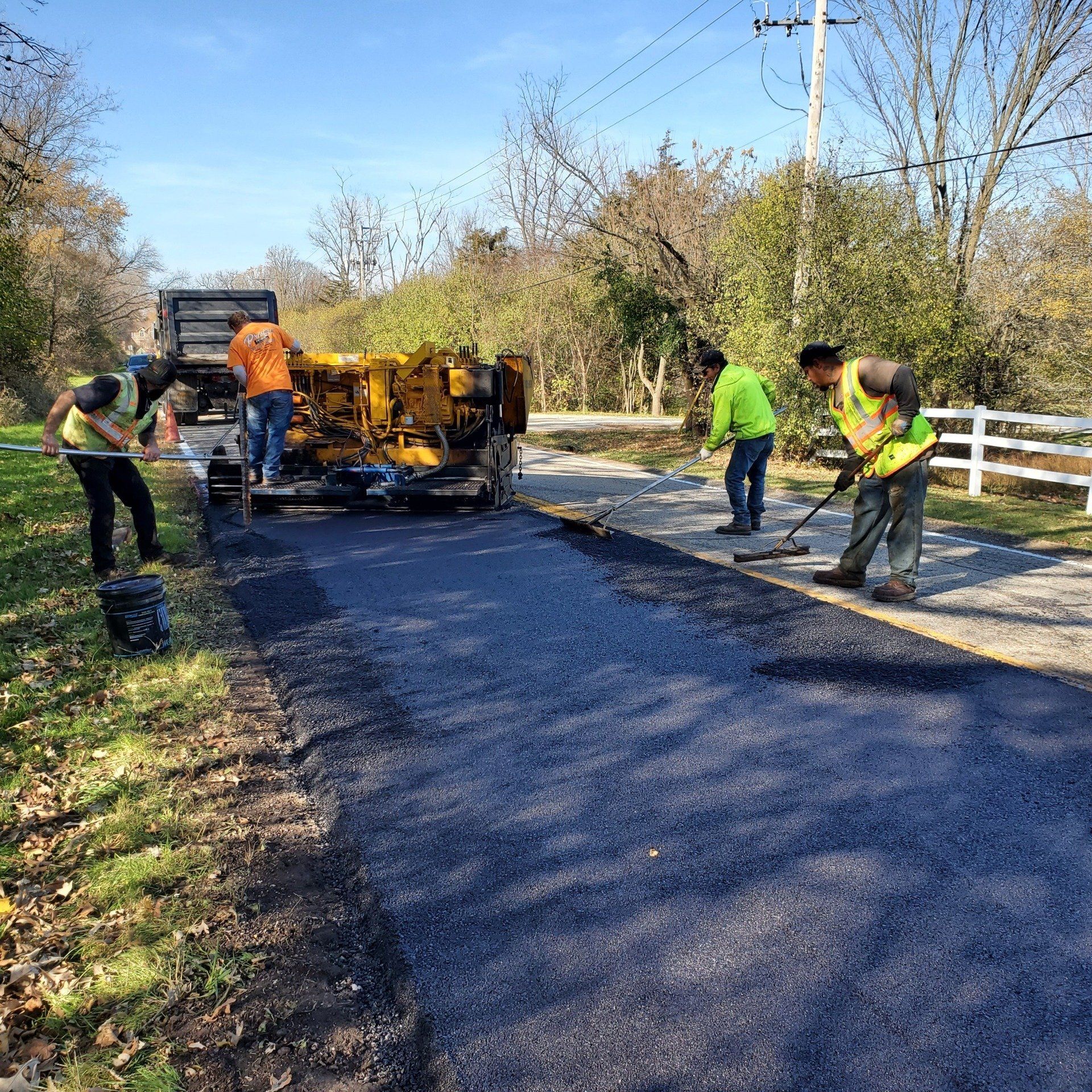 Four construction workers wearing safety vests work together to pave a residential driveway with fresh black asphalt.