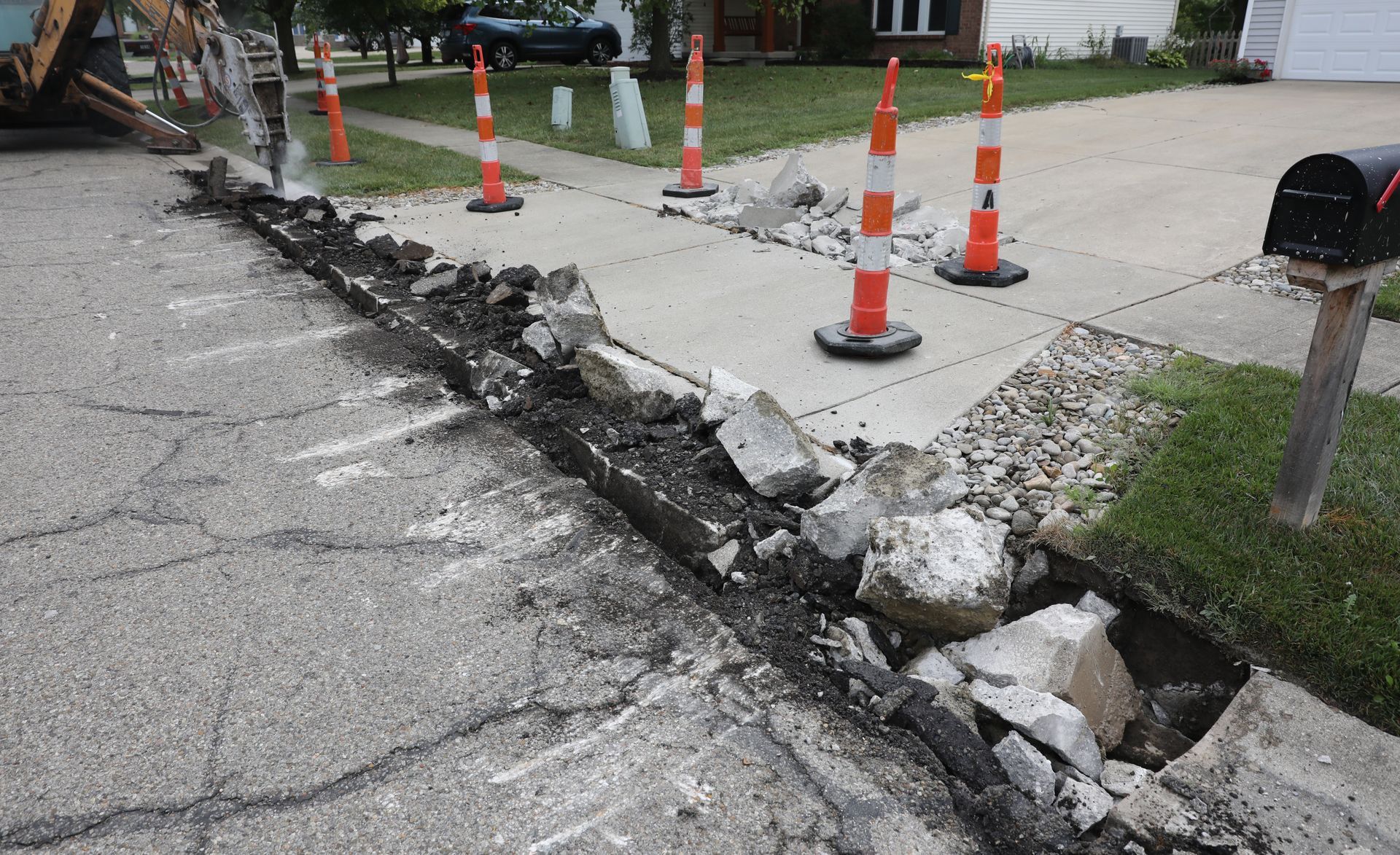 Excavator demolishing a cracked driveway, exposing broken concrete, gravel base, and safety cones. Excavator demolishing a cracked driveway, exposing broken concrete, gravel base, and safety cones.