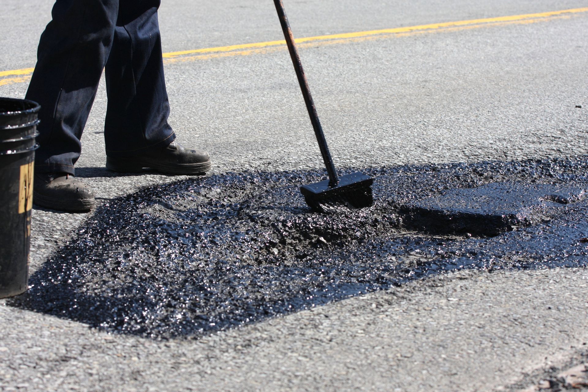 A road construction worker spreads tar in a pothole. Person patching asphalt on a road with a brush, black material visible.