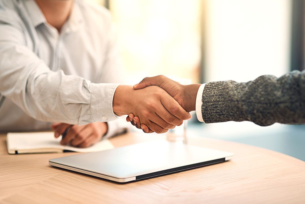 Two people shaking hands over a table with a laptop and notebook.