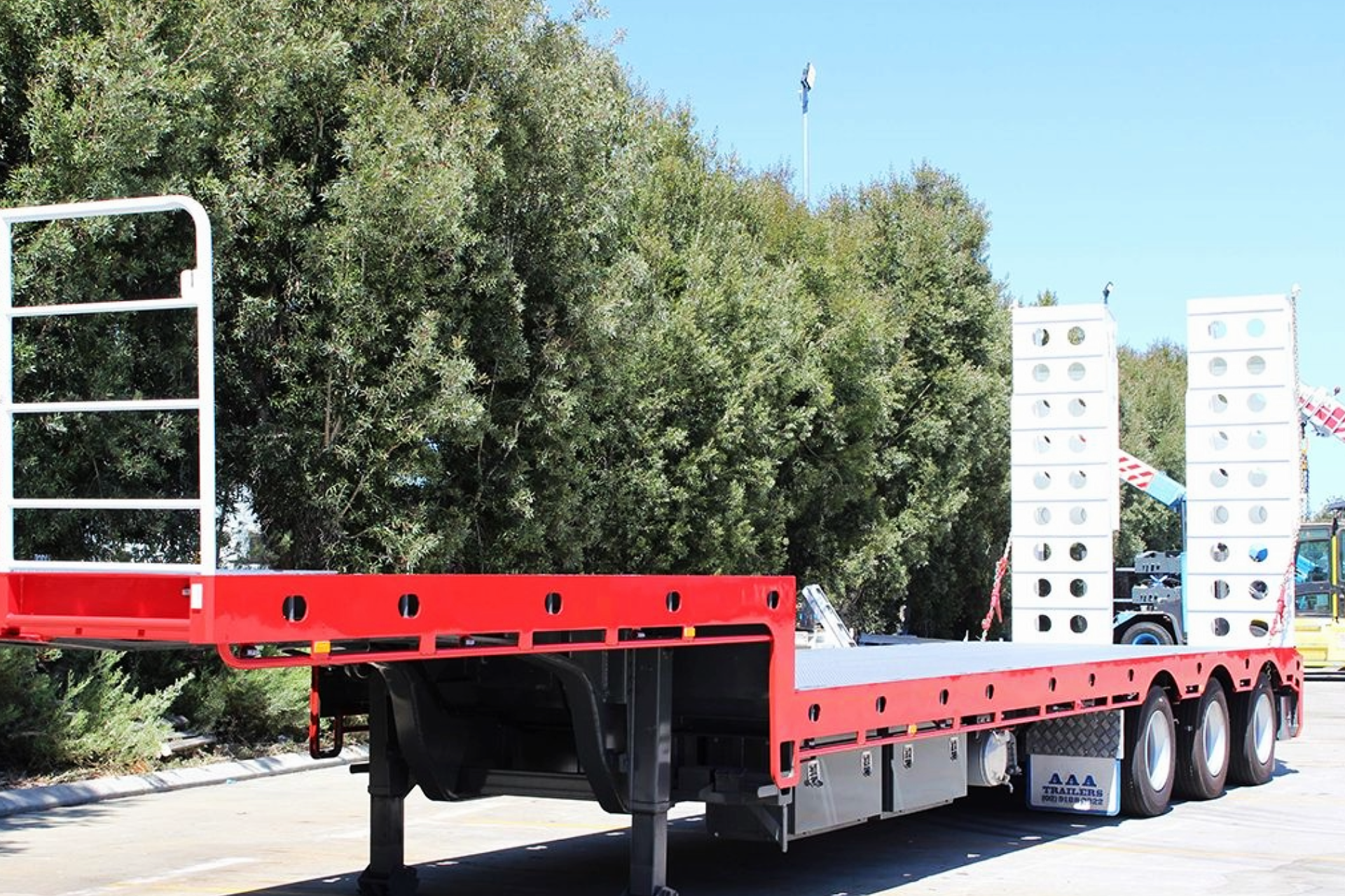 Red flatbed trailer with ramps parked outdoors beside trees on a sunny day