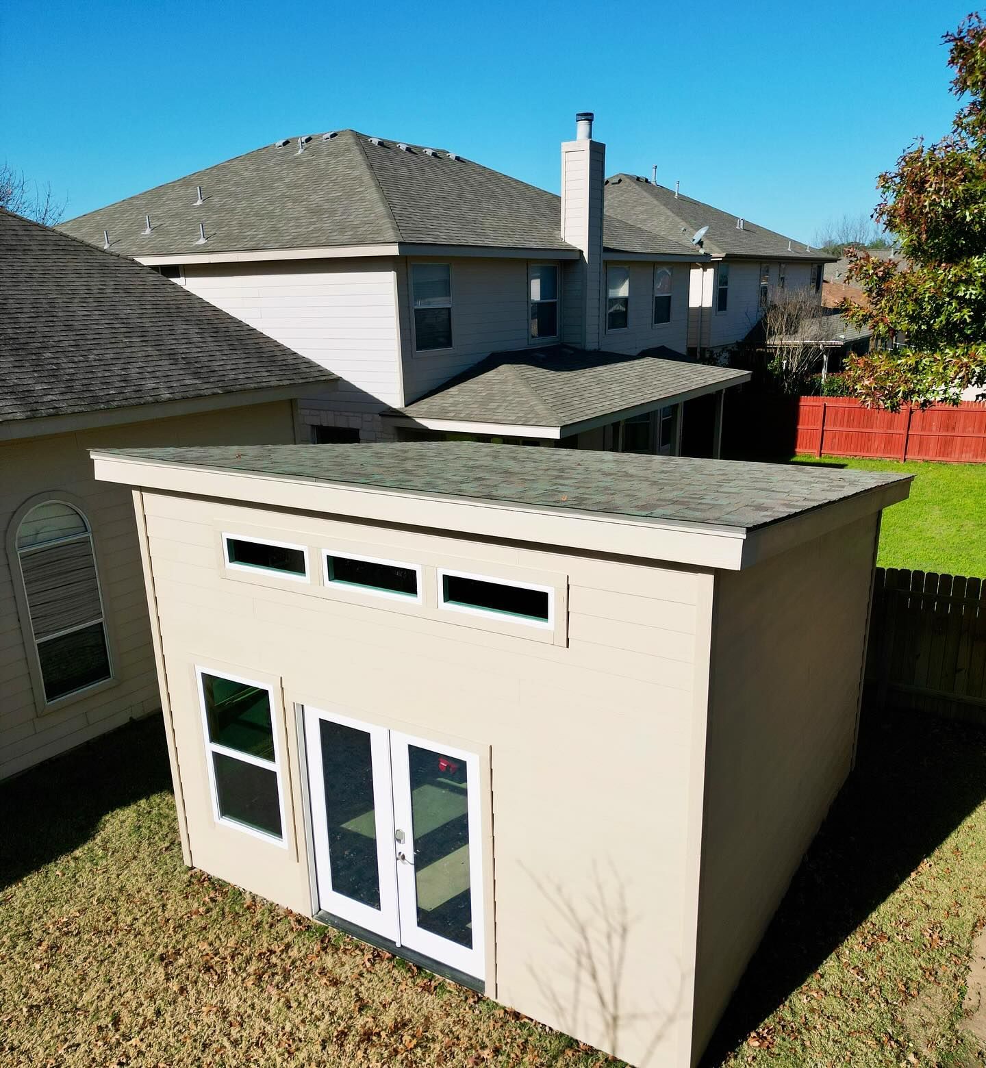 Small beige backyard shed with white French doors and windows in a fenced suburban yard