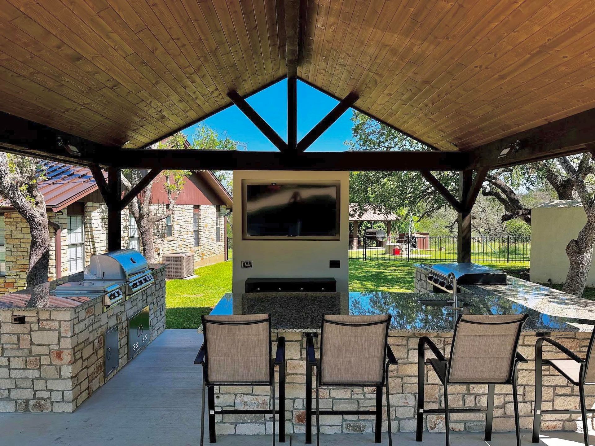Covered patio with TV, bar seating, and stone grill beside a grassy yard