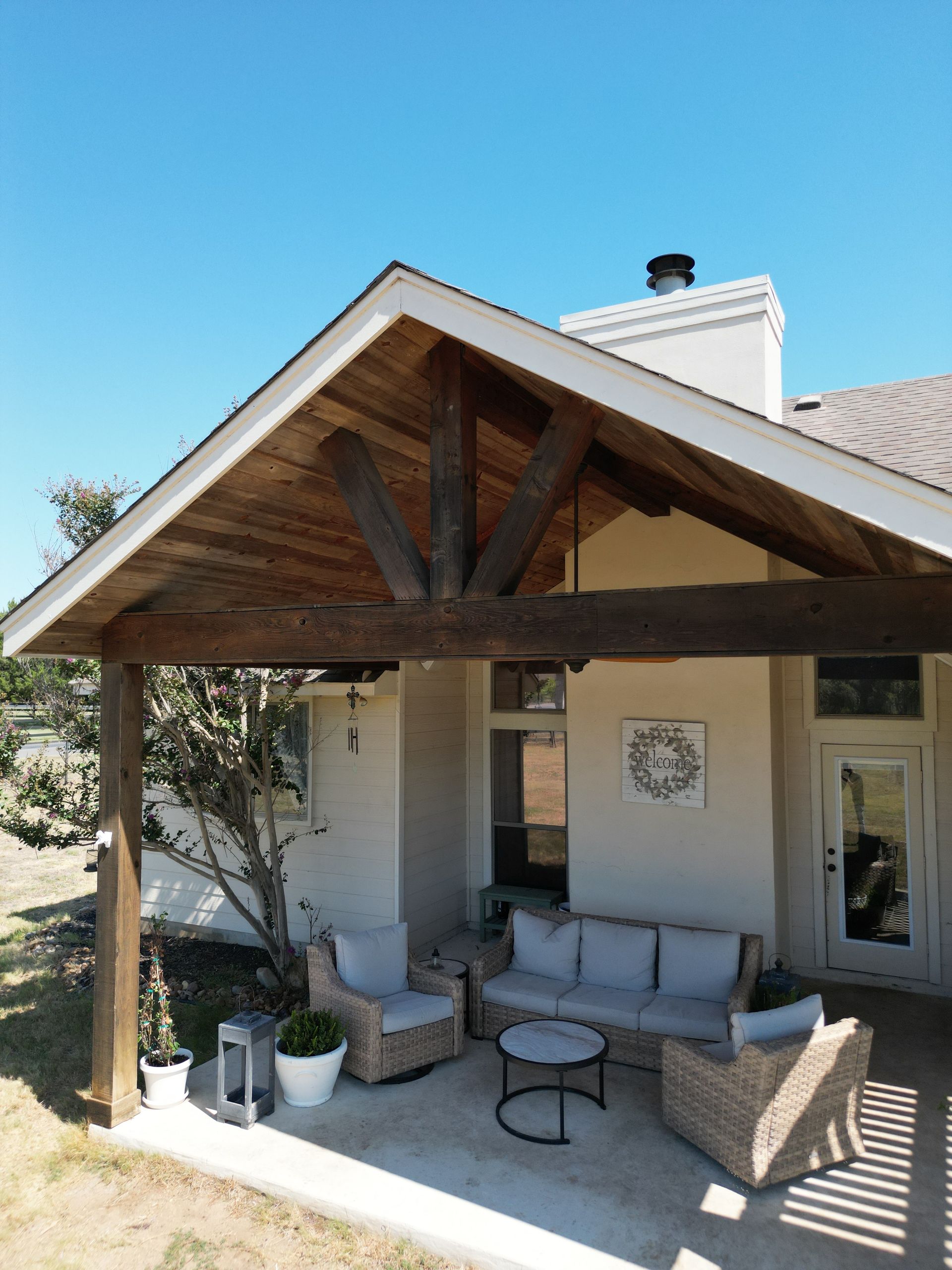 Covered patio with wooden beam roof, wicker seating, and potted plants outside a house.