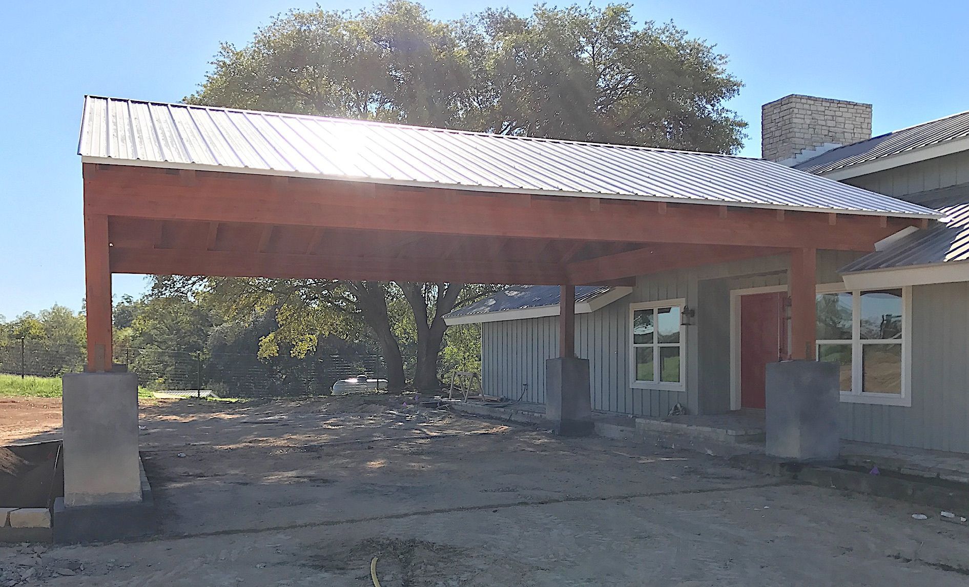 Small house with a red metal carport roof and dirt yard, with trees in the background