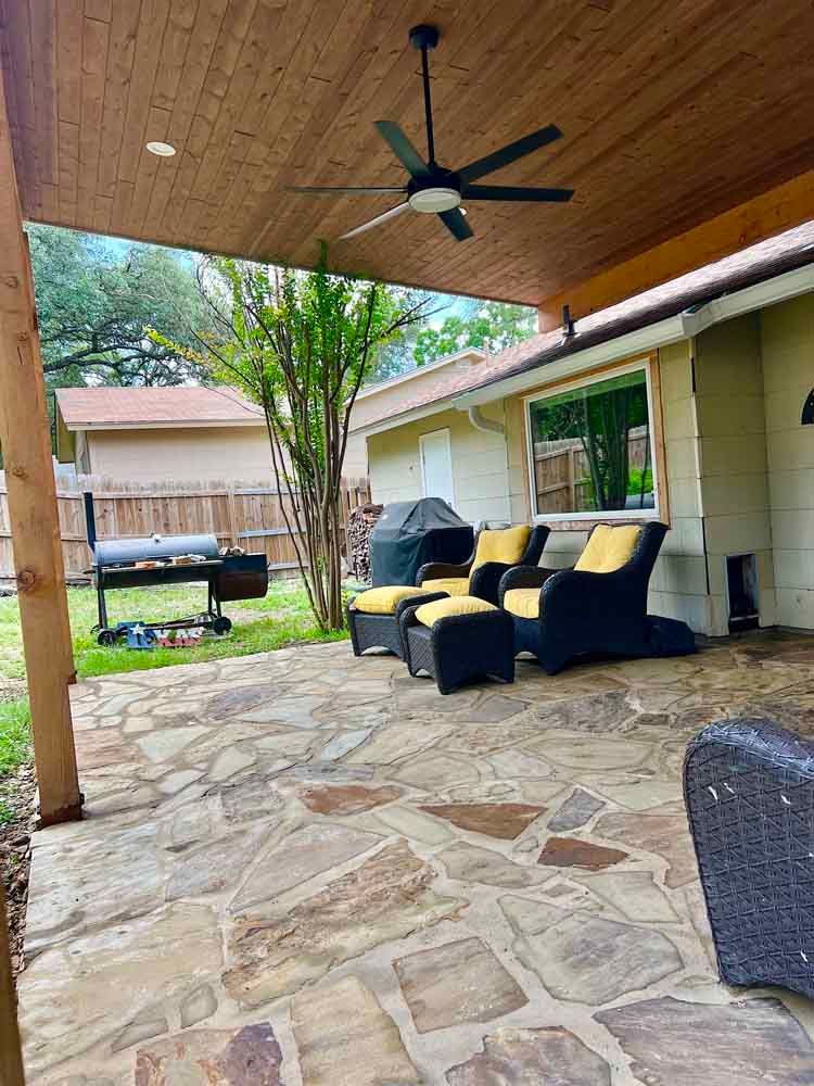 Covered patio with stone floor, ceiling fan, wicker chairs, grill, and a view of the backyard.