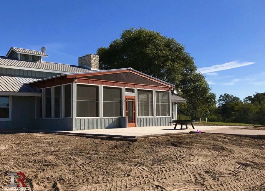 Single-story house with screened porch and red roof, beside a sandy yard under a blue sky.