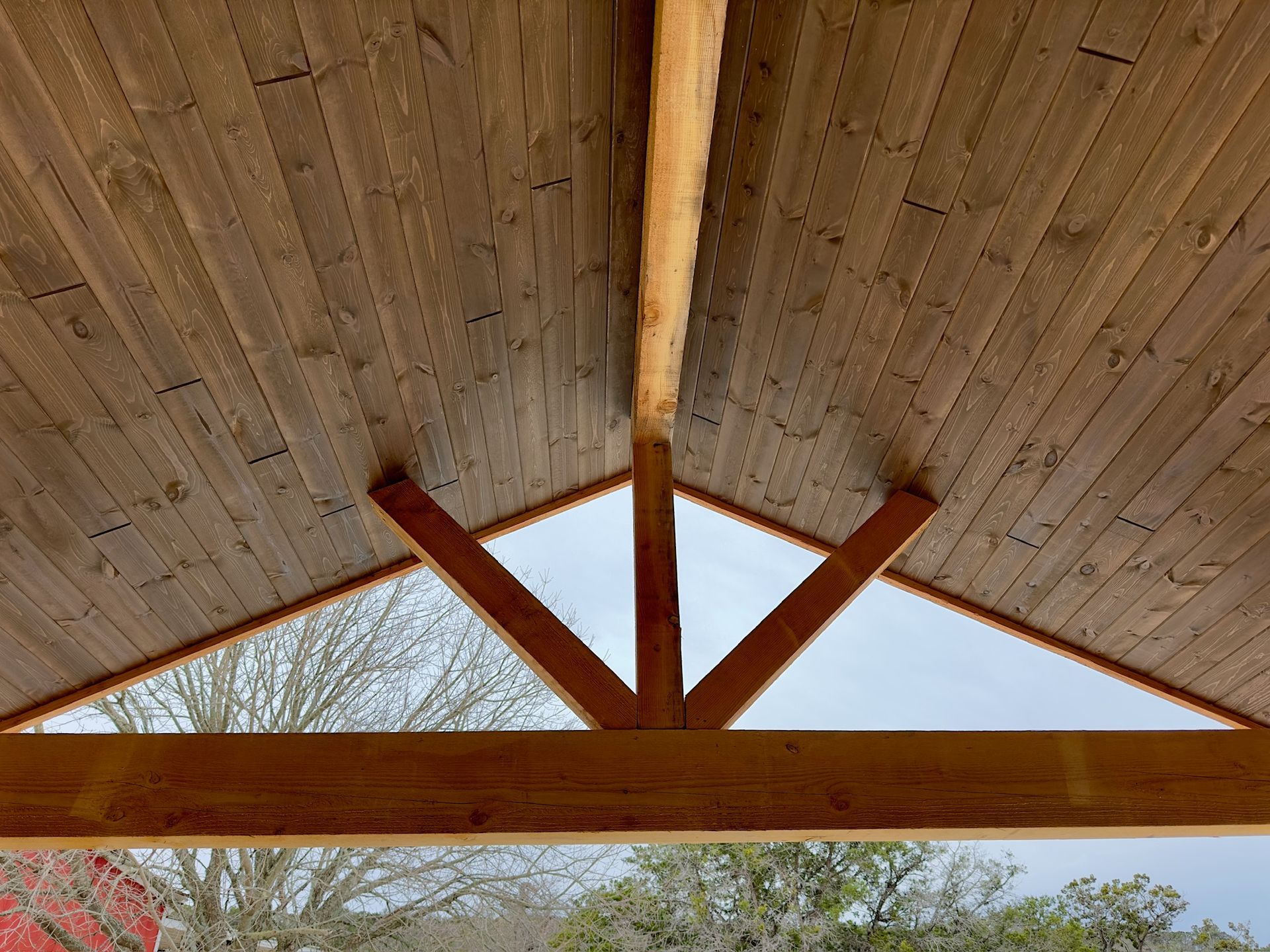 Wooden pavilion ceiling with exposed beams and a view of trees outside.