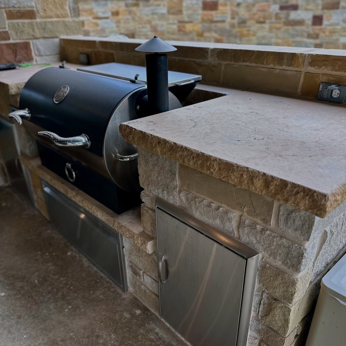 Black outdoor smoker grill built into a stone counter beside a brick wall