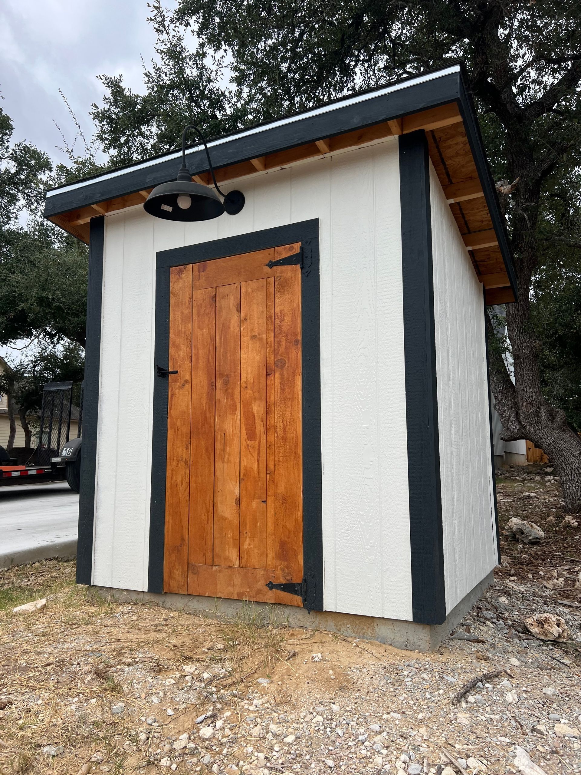 Small white utility shed with dark trim, a wooden door, and a slanted roof on a gravel lot.