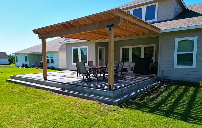 Covered backyard patio with outdoor dining table, chairs, and a lush green lawn beside a house.