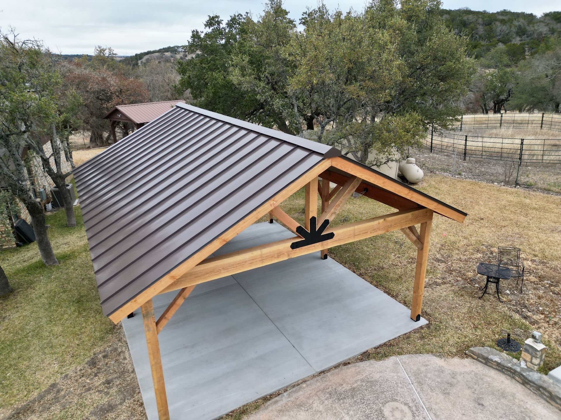 Wooden carport with a gray metal roof over a concrete pad in a rural yard