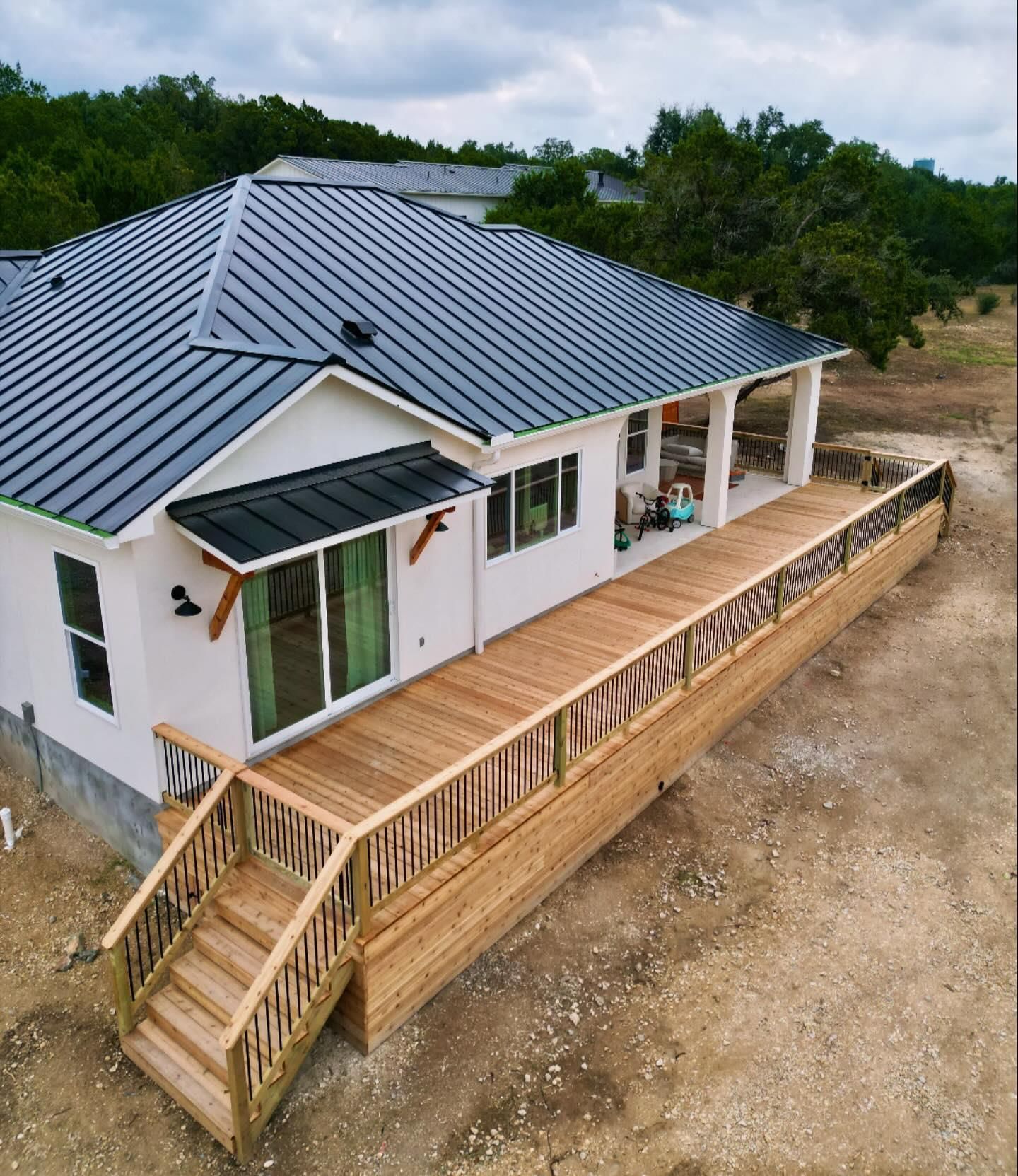 Modern white house with gray roof and long wooden deck on a dirt lot, surrounded by trees