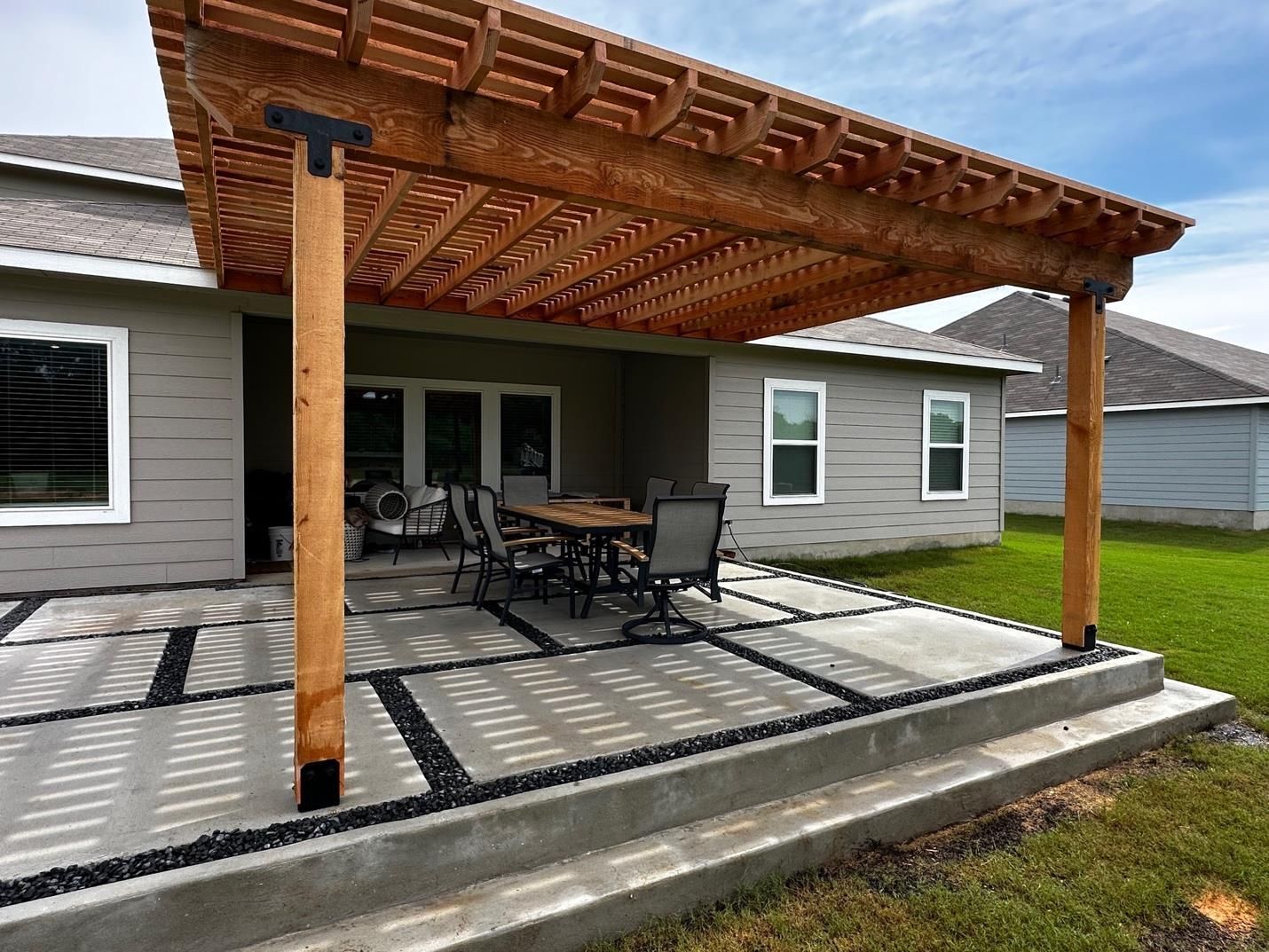 Wooden pergola over a patio with outdoor dining furniture beside a gray house