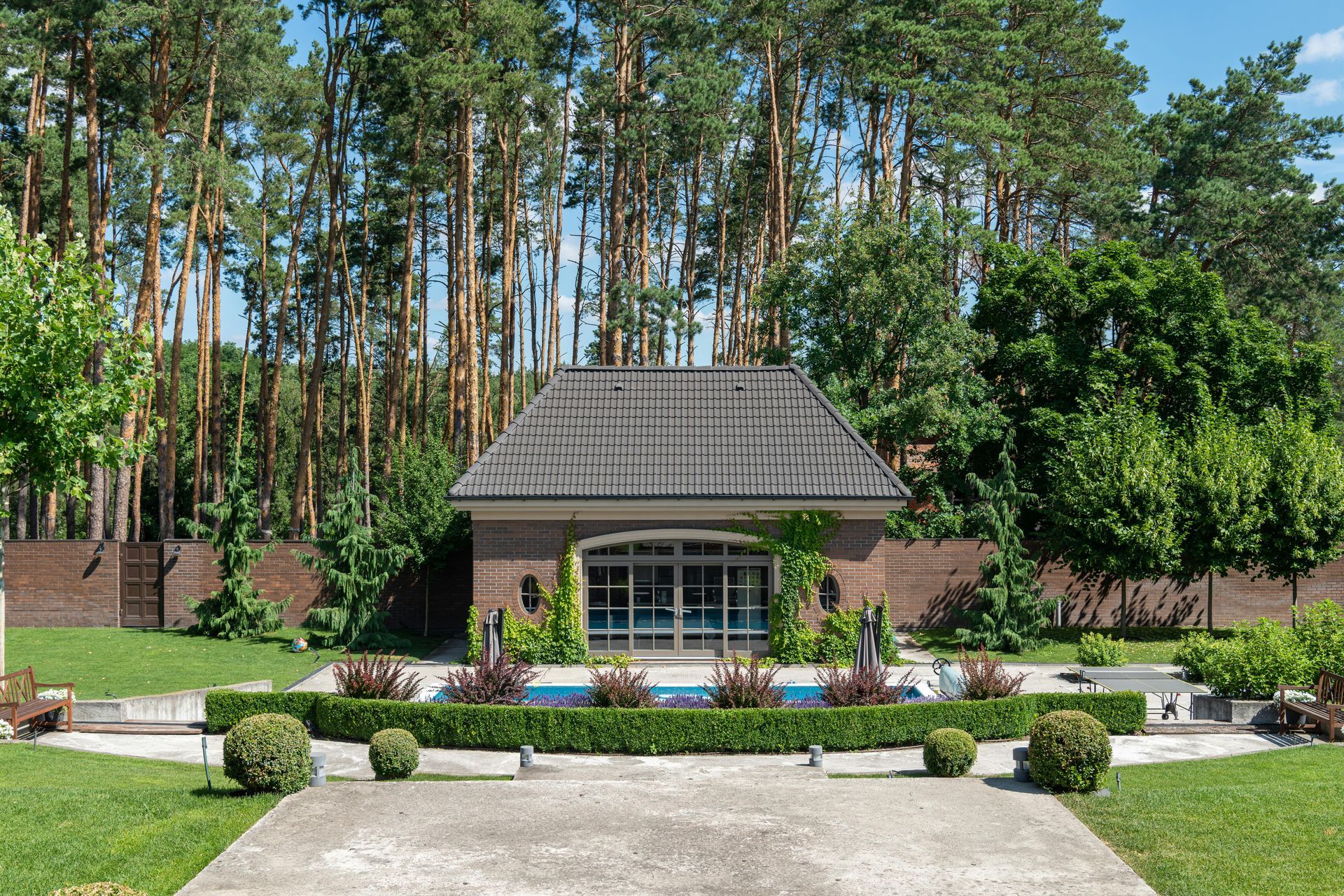 Garden pavilion with a shingled roof, flower beds, and tall trees in the background