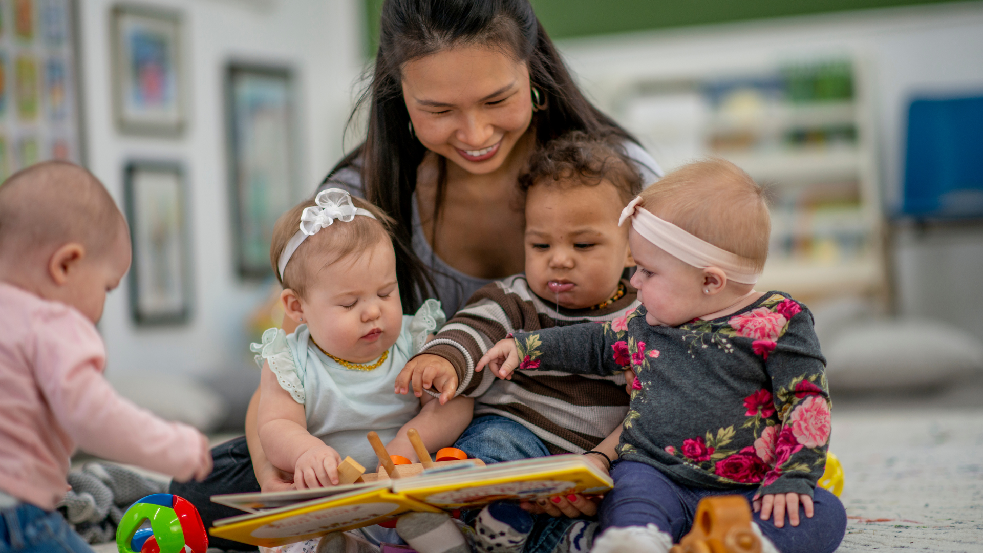 Woman reading a book to four toddlers seated on a carpet in a bright playroom