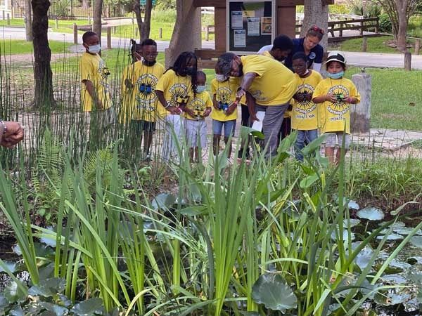 Children in yellow shirts standing near pond plants in a park, some wearing masks