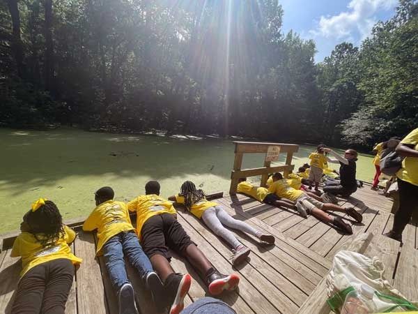 People in yellow shirts lie on a wooden dock beside a green pond in a forest clearing.
