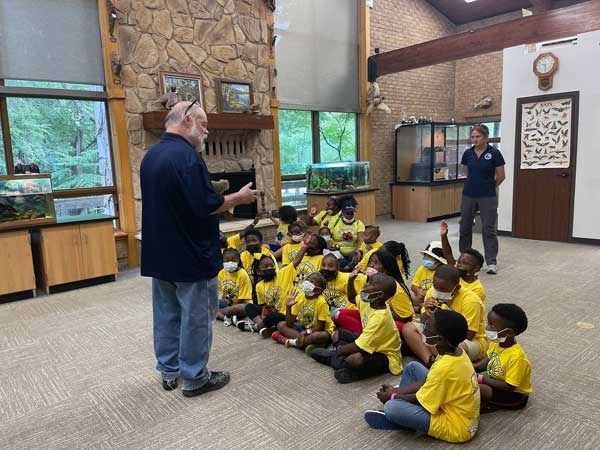 Speaker addressing seated children in yellow shirts inside a community hall