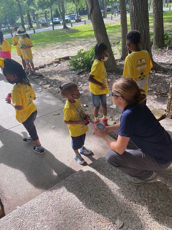 Children in yellow shirts standing with an adult outdoors in a park, one child receiving a snack