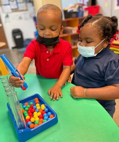 Two children wearing masks play a color-matching game with colorful balls at a table.