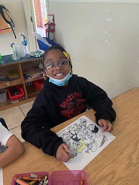 Child smiling at a classroom desk, holding a coloring page with a mask under the chin.