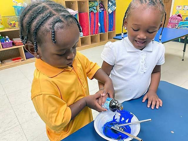 Two children in a classroom pour blue paint into a bowl on a blue table