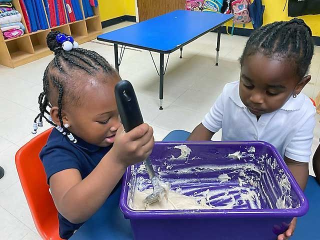 Two children mixing dough in a purple bin in a classroom, one holding a spoon and smiling.