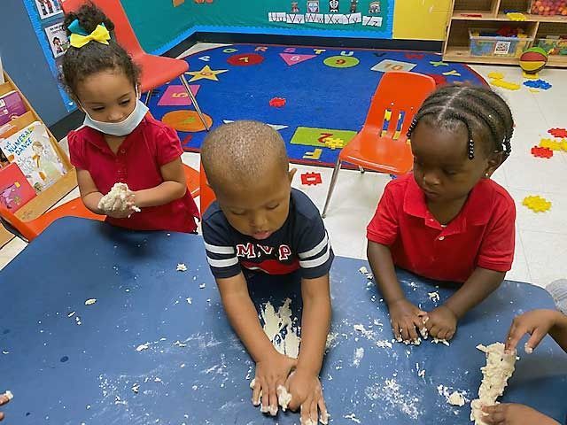 Three children in red and navy shirts playing with white sand on a classroom floor, with colorful chairs nearby