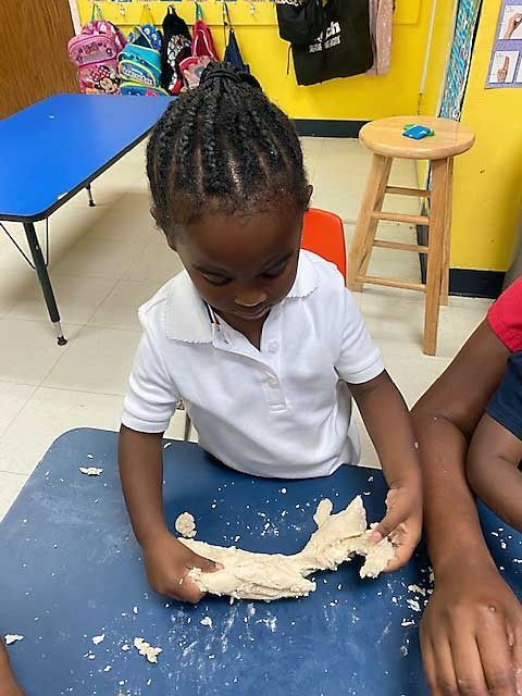 Child kneading dough at a table in a classroom, with flour scattered on a blue mat.