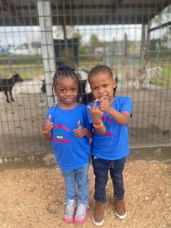 Two children in blue shirts standing in front of a wire fence, smiling and holding snacks