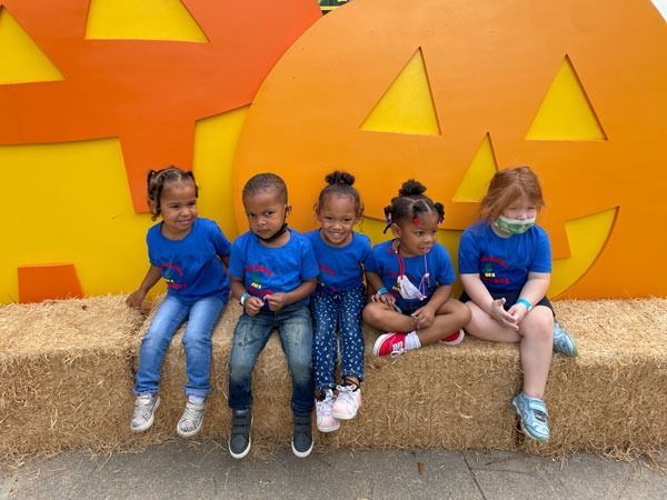 Five children sit on hay bales in front of a yellow fall pumpkin display.