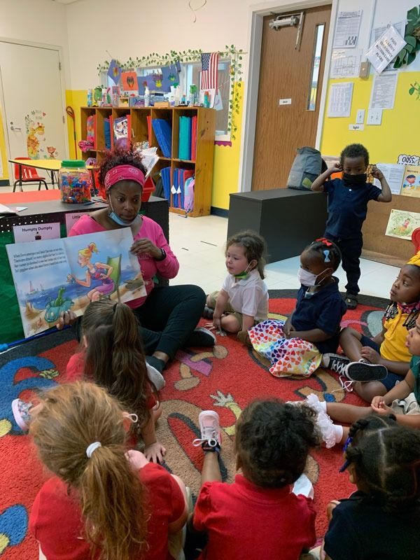 Children sitting on a classroom rug, listening to a teacher reading a colorful book.