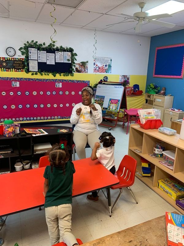 Teacher standing by red table in colorful classroom with two children facing her and shelves of supplies.