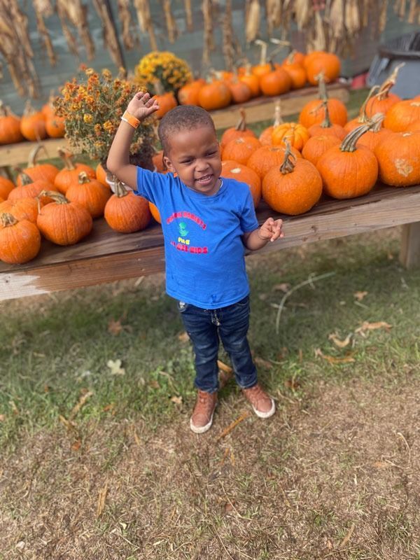 Child in blue shirt standing by pumpkins at a fall display, raising one arm and smiling
