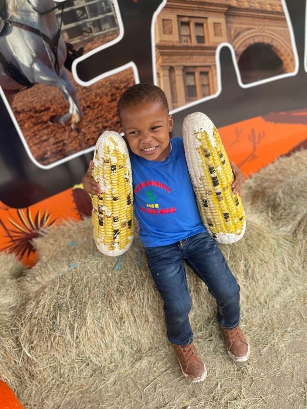Smiling child sitting on hay bales at a harvest display, holding two yellow corn cobs.
