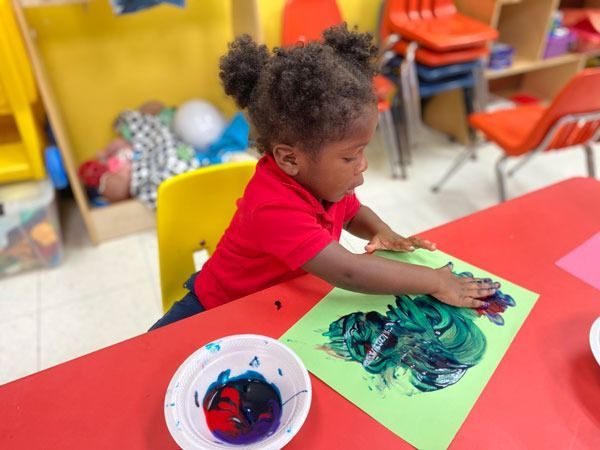 Child painting with hands on green paper at a classroom table, with a paint bowl nearby.